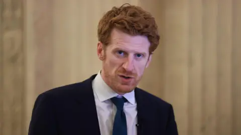 PA Media SDLP MLA Matthew O'Toole, with short red hair, wearing a white shirt, navy tie and navy suit. 