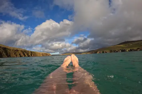 Emma Williamson Two feet sticking out of the sea, in a selfie taken by a wild swimmer, with land and clouds in the distance.
