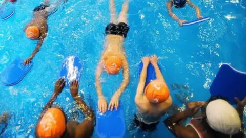 A number of people swimming in a swimming pool, while wearing swimming caps and holding blue floats.