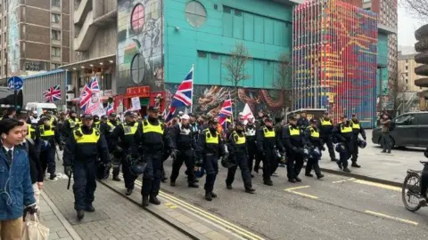 Several rows of police in black uniforms and hi-vis jackets walk ahead of protesters carrying Union Jack flags in Bristol city centre.