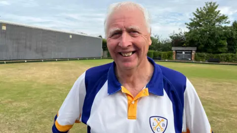 Ollie Conopo/BBC Alan Simms, looking straight at the camera and smiling, with short grey hair, he is standing on a bowling green and is wearing a white top with blue and gold on it. 