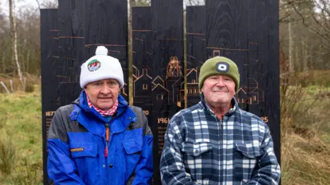 West Cumbria Rivers Trust Former miners Joseph Ritson and David Cradduck stand in front of black wooden installations in a woodland. Joseph is wearing a blue waterproof jacket and a white woolly hat while Dave wears a black and white tartan fleece jacket and a green woolly hat.
