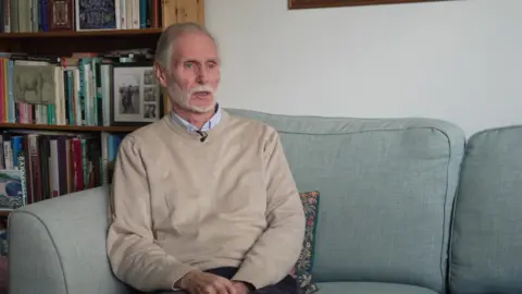 Jim with white hair and a white beard, wearing a beige jumper over a blue collared shirt, sits on a pale green sofa in front of a full bookcase as he speaks during an interview