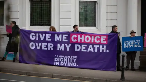 PA Media Dignity in Dying campaigners stand in front of a white building with large windows, holding a large purple sign, with white lettering that reads: "give me choice over my death".