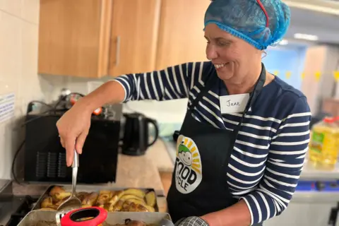 Jon Wright/BBC Volunteer cook Jane Hunt turns over some potato wedges in a pan. She is wearing a branded FoodCycle apron and a blue hair net