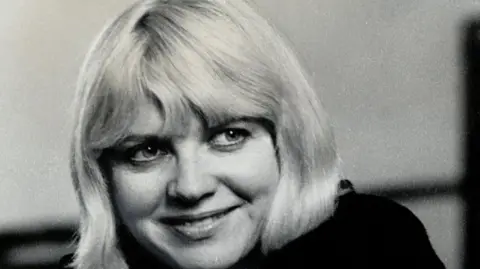 Getty Images A black-and-white close-up shot of a woman in her mid-30s, with short, bobbed blonde hair, wearing a black top, smiling and looking away to the right of frame.