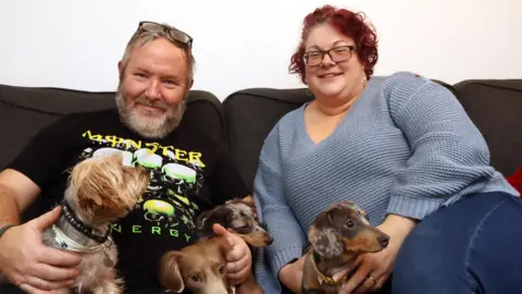 SUPPLIED Man and woman sat on a dark brown sofa infront of white wall with four dogs on their laps