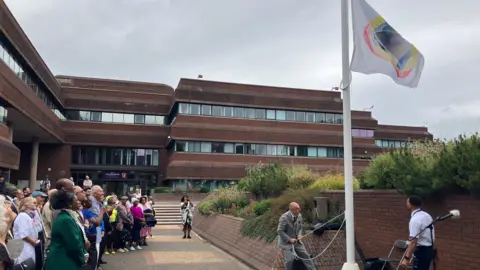 Dignitaries and members of the public gather as the Windrush flag is raised outside the Civic Centre. Lines of people are standing in front of a white flagpole with the colourful flag waving in the breeze.