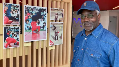 A man wearing a blue denim shirt and blue cap standing next to colour photographs on a wooden slatted display wall. 