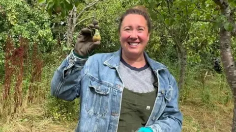 BBC Victoria Holden has dark hair tied back in a bun. She is wearing a blue and white striped t-shirt, green dungarees, a denim jacket and blue gloves. She is stood in an orchard and is holding up a small pear to the camera.