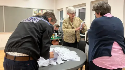 Four people, including a police officer, stand around a table which has ballot papers on it for counting at the 2026 Jersey election.