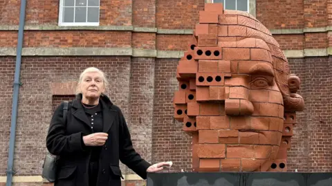 A woman with grey hair, wearing a black coat over a black cardigan, is standing next to a brick-built sculpture showing the partial face of Josiah Wedgwood with a building behind her filling the rest of the backdrop.