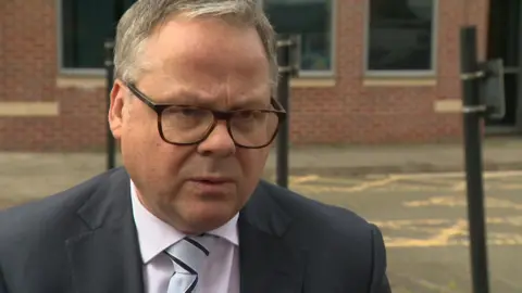 A man with short greying hair. He is wearing glasses and a dark suit and tie. He is standing outside a brick office building.