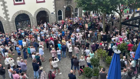 Large crowd gathered in a town square between historic buildings and trees, with people standing closely together around a small group of speakers in the centre.