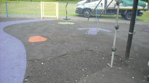 Black rubber surfacing with blue and orange spots next to play equipment. A large hole is in the floor. 
