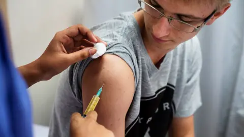 A young man aged about 19 wearing a grey t-shirt and glasses recieving a vaccine in the top of his arm 