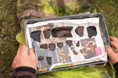 PA Media A pair of hands holds a tray containing broken pieces of ancient pottery