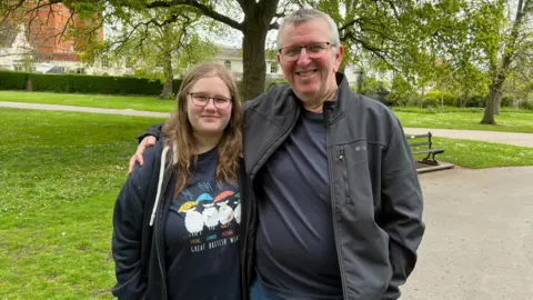 A young woman with blonde hair and glasses stands next to her father who has grey hair and glasses in a park.