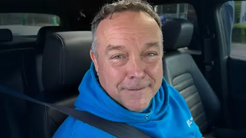 Close-up of a person wearing a blue hoodie sitting in the front seat of a car with a seatbelt on.