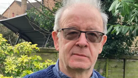Peter Anderson Peter Anderson looks at the camera. He is wearing dark glasses and a blue shirt and jumper on top. He is standing in a garden with green foliage behind him. He has white, short hair. 