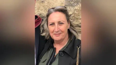 Family handout A woman with short grey hair wearing a black jacket, khaki green shirt, and a bag with a gold chain. She is smiling at the camera and has a pair of sunglasses on her head.