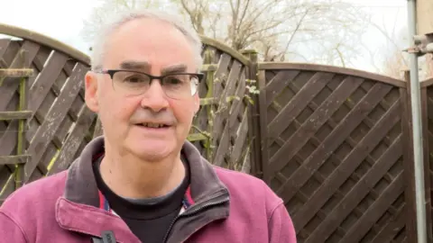 John Hall stands in front of brown fencing in his front garden. He is wearing a purple zip up jacket and glasses.