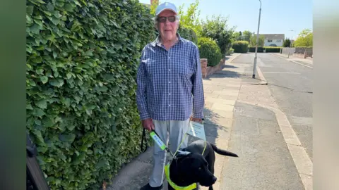 Mark Denten/BBC A close-up photo of David, who is wearing a blue chequered shirt, a light blue cap, and dark-tinted glasses. He holds the lead of his sight dog, which is wearing a luminous yellow harness.