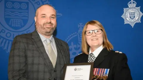 Simon Kirk accepting an award from a police officer with a blue backdrop 