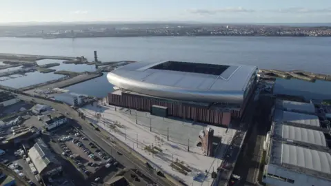 BBC An aerial view of Everton's new stadium, a curved silver structure on dockland with the River Mersey in the backdrop.