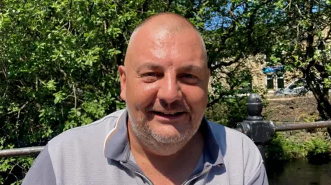 A bald man wearing a grey polo shirt with a zip-up front sitting outside with railings in the background.