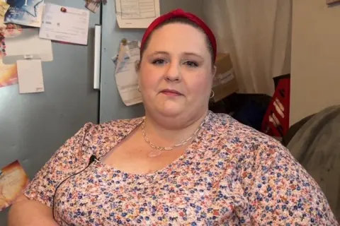 Tenant Karen Bray is seated, wearing a floral dress and silver necklace. She has long brown hair tied back in a headband. She is pictured on her sofa, directly next to her fridge. 