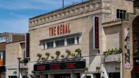 Mike Kemp/In Pictures via Getty Images An art deco building constructed of Bath stone glows in the sunshine. It is a former cinema with a board on its front that says "The Regal", with large red letters above that also say "The Regal". Some foliage is being grown in pots on the front.