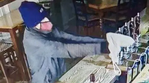 A blurred CCTV image of a man wearing a blue beanie and red face mask, pointing a toy gun concealed in a tote bag at the victim who is standing behind a bar in a hotel.