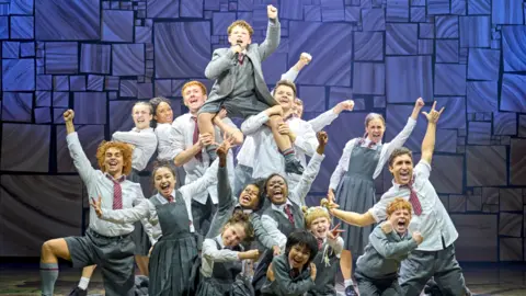 Matilda the Musical The cast of Matilda the Musical are huddled together in the final pose of a big group number. Some older cast members are holding children up in the air, with other kids gathered at the front with their fists pumped to the sky or holding out peace signs. 