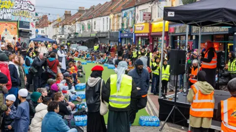 A crowd of people fill a street in Bristol. There is a long green runway in the middle of the street and a stage on the pavement. Some of the people are wearing high vis jackets.