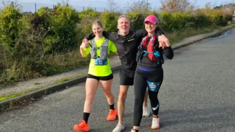 The Rehab Physio, Bromborough Mark running along a road towards the end of the half marathon - he's now being supported by two women with his arms around their shoulders, and the three of them are running together. All are wearing running kit.