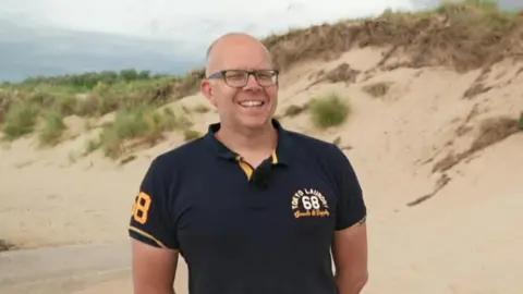 Man in a navy-blue polo shirt stands on sand dunes smiling. He is bald and wears glasses.