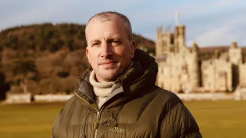 Dr Langlands wears a brown coat and stands in a field with a mountain and building in the background, he looks at the camera with a closed-mouth smile.