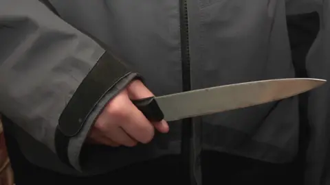 BBC The photo shows a person's hand holding a large knife. The knife has a black plastic handle. The wielder of the weapon wears a gray waterproof coat.