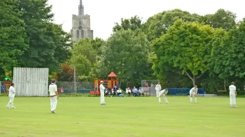 Andy Lloyd/BBC A village cricket match in Ashwell, Hertfordshire, on a summer's day in 2008. Cricketers in their whites are playing on a pitch in front of a church and are being watched by spectators.