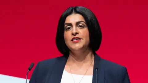 PA Media Home Secretary Shabana Mahmood is in a white top and a navy blue jacket as she delivers a speech during the Labour Party conference. Her hair is cropped into a neat bob, and she wears red lipstick.