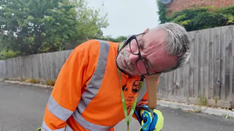 A man wearing glasses and a fluorescent orange top places his ear against the wooden end of a metal listening stick as he tries to find a water leak