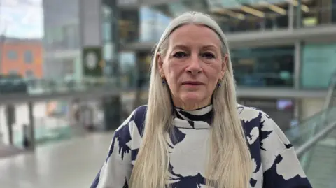 Paul Moseley/BBC A woman with long light-coloured hair stands indoors in a spacious, modern atrium. She's wearing a patterned sweater and faces the camera. Behind her, glass railings, escalators, and multi-level walkways are visible. 
