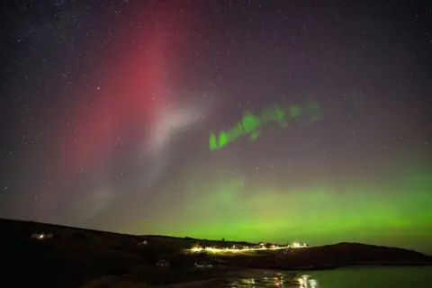 Gary Macleod The Northern Lights in the north coast of Scotland. There are green and red lights across the sky with a village below. 