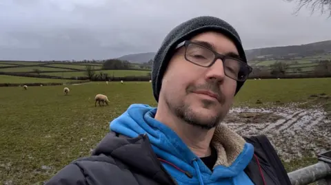 Dean Cooper Deen Cooper is seen in a black jacket, blue jumper and beanie. He is taking a selfie in front of green fields. The sky is grey.