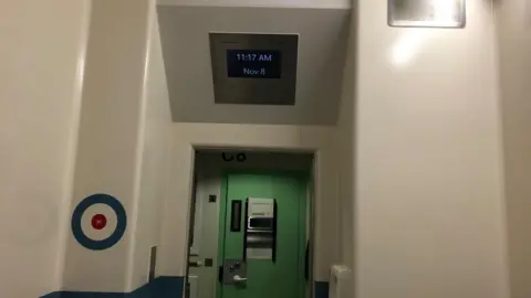 Vikki Irwin/BBC A view of a green prison door looking out from the cell with a TV screen hanging above it. There is also a target on the walls. The image illustrates how in-cell technology is used in police cells.  