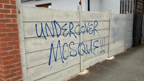 Jami Community and Education Centre A white fence around a building has blue spray painted graffiti that reads "undercover mosque". More graffiti next to it is blurred out