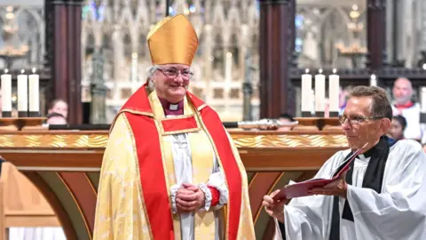 Diocese of Ely The Right Reverend Sarah Clark has white hair in a ponytail. She is wearing red and clear framed glasses and clerical clothing. She is standing inside a cathedral. 
