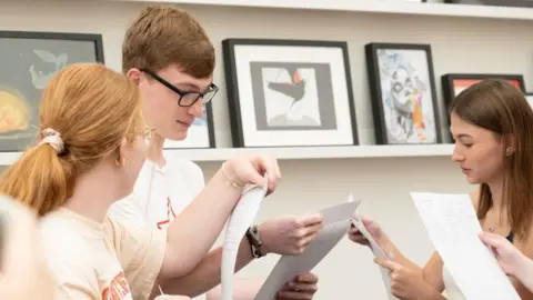 Aureus School Three teenagers in white tops read their results from pieces of paper in a candid shot. 