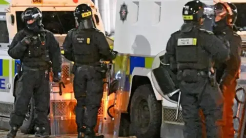 Two police Land Rovers in the background with four police officers in helmets standing in front of them. They are holding riot shields.